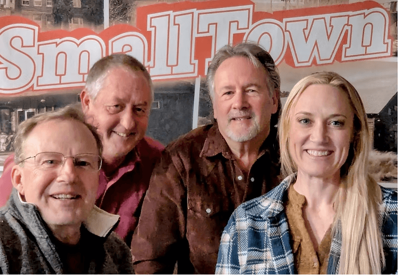 Four smiling friends posing together in front of a 'Small Town' banner in Central Oregon.
