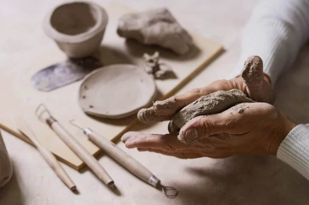 Hands kneading clay into a ball, surrounded by pottery tools and a partially formed clay bowl in Central Oregon.