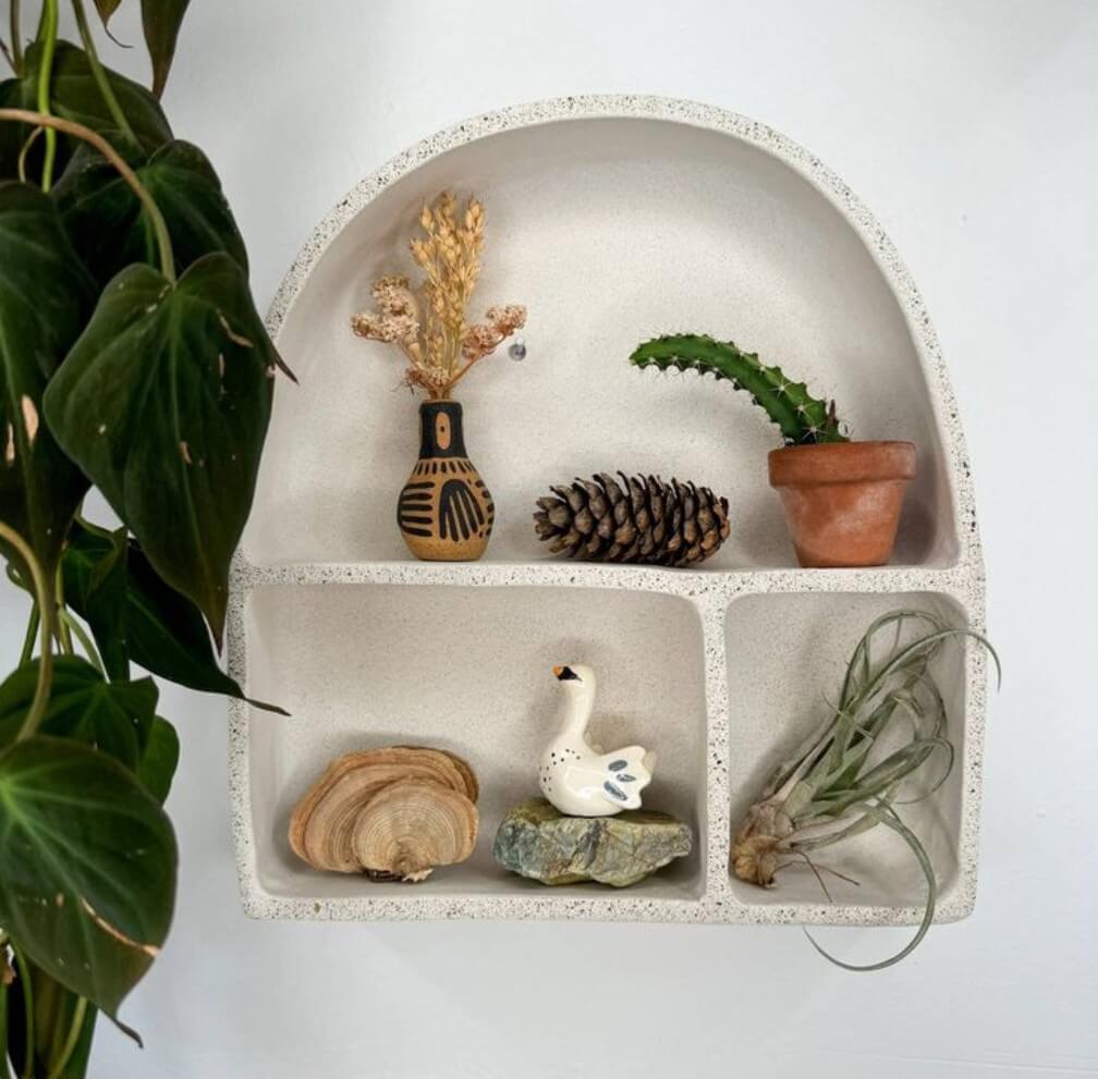 A white arched wall shelf displays a variety of items including a cactus in a terracotta pot, a wooden duck figurine, dried flowers in a black and white vase, and a pine cone alongside other natural elements in Central Oregon.