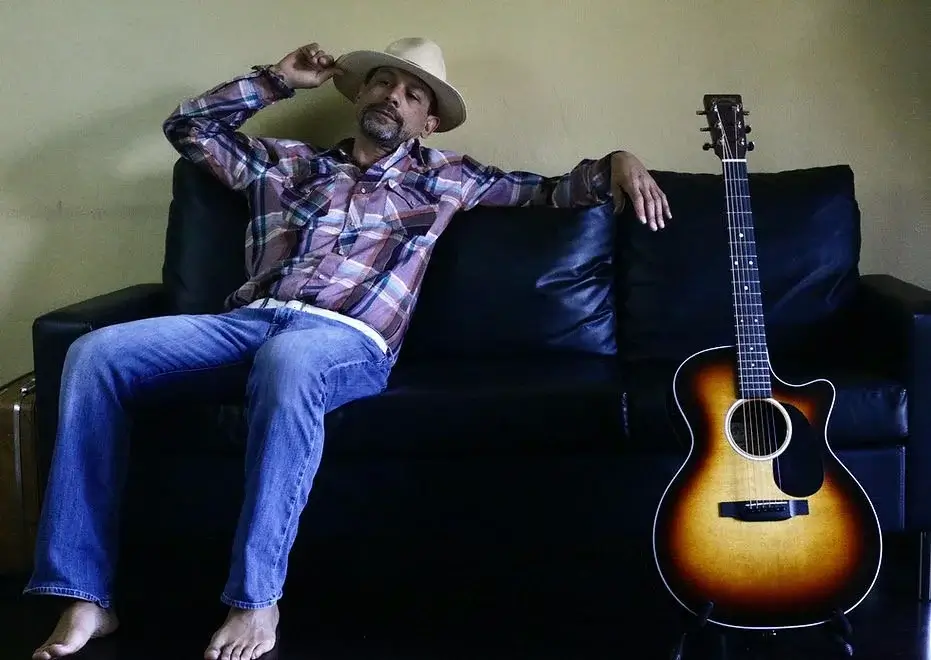 A man wearing a cowboy hat and plaid shirt relaxes on a black leather couch with an acoustic guitar beside him in Central Oregon.