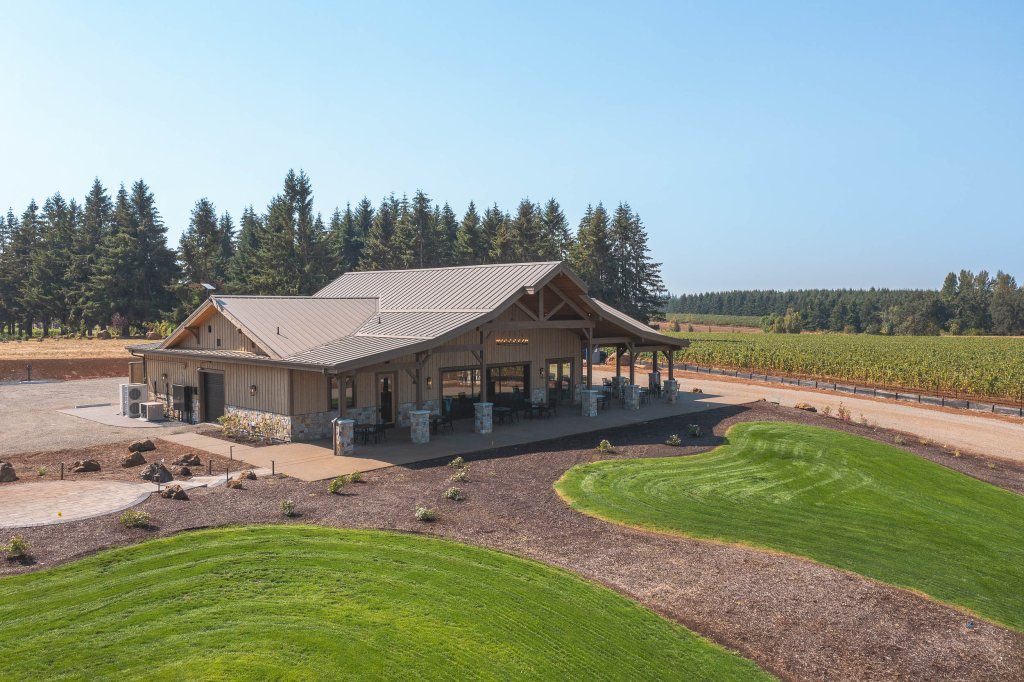 A rustic wooden building with a metal roof sits amidst a lush green lawn and a vineyard, surrounded by tall evergreen trees under a clear blue sky in Central Oregon.