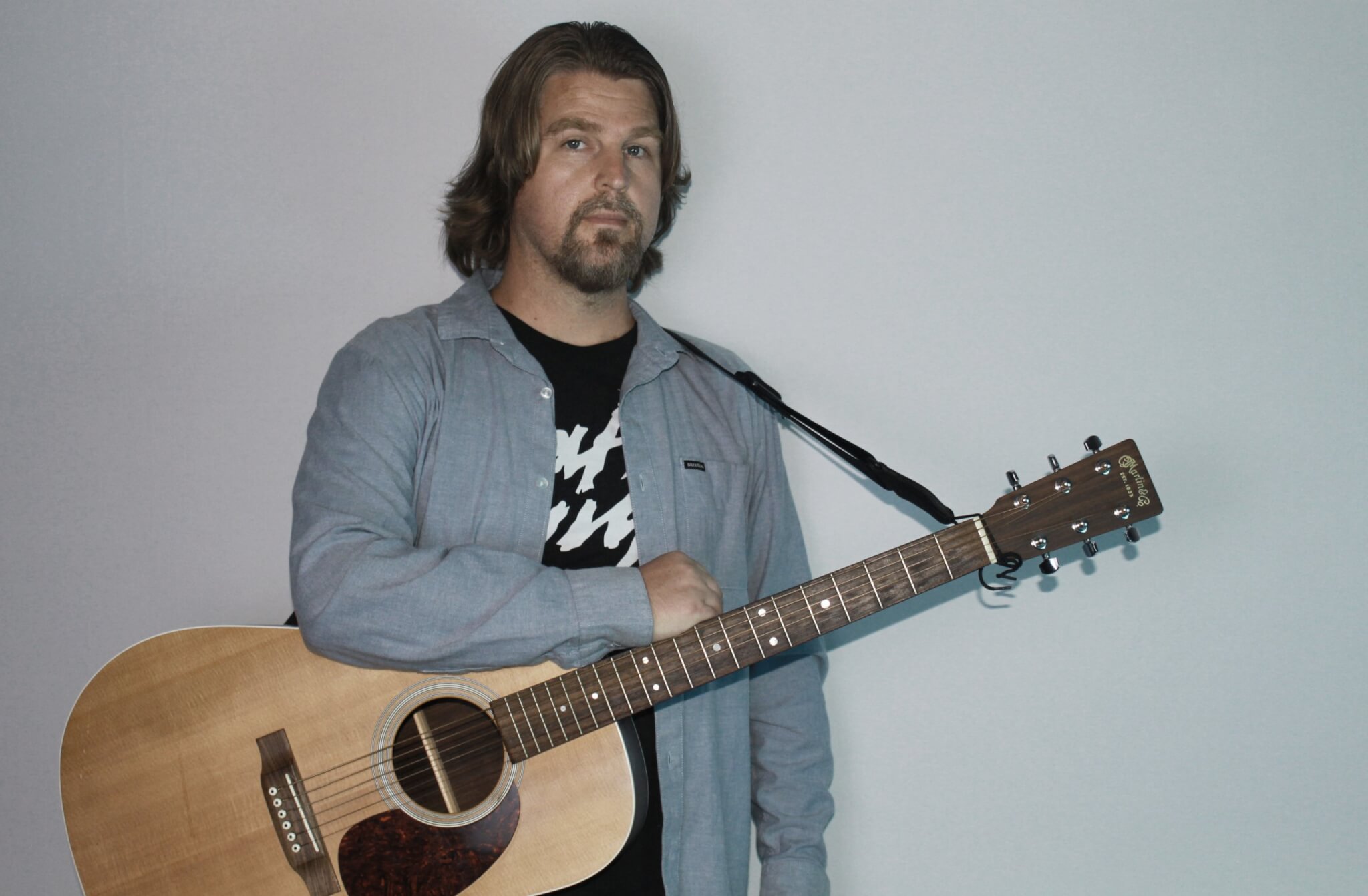 A man with long hair and a beard wearing a blue shirt over a black graphic tee holds an acoustic guitar against a white wall in Central Oregon.