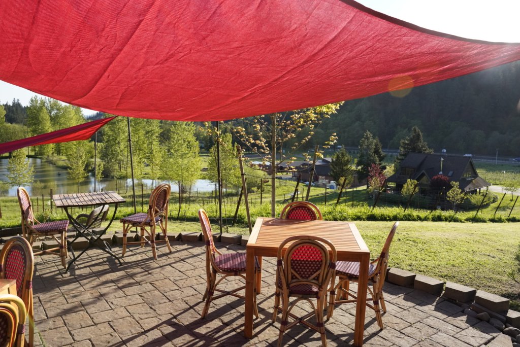 A serene outdoor dining area with a red canopy, wooden tables and chairs, and a view of a tranquil lake surrounded by lush greenery and distant mountains in Central Oregon.