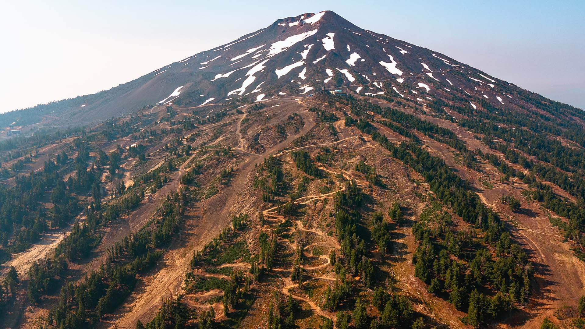 A majestic snow-capped mountain peak rises above a rugged landscape dotted with coniferous forests and winding dirt trails in Central Oregon.