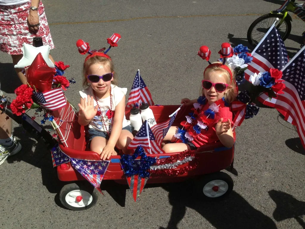 Two young girls dressed in patriotic attire wave from a decorated red wagon during a Fourth of July parade in Central Oregon.