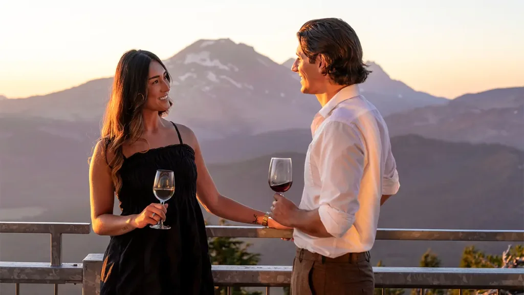 A couple enjoys a romantic sunset on a mountain peak, sipping wine and gazing at the snow-capped peaks and valley below in Central Oregon.