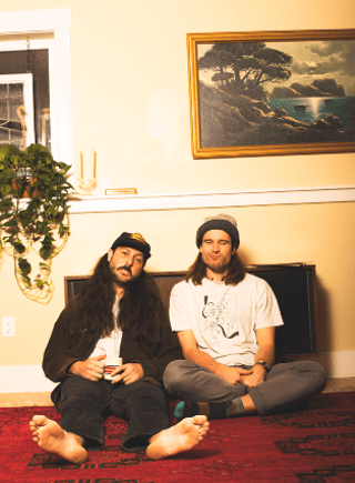 Two men with long hair and beards sit cross-legged on a red rug, one wearing a cap and the other a beanie, with a framed landscape painting and a hanging plant in the background in Central Oregon.