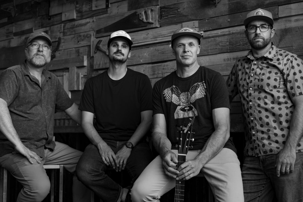 Four men in casual attire, including hats and glasses, pose together against a rustic wooden wall, with one holding an acoustic guitar in Central Oregon.