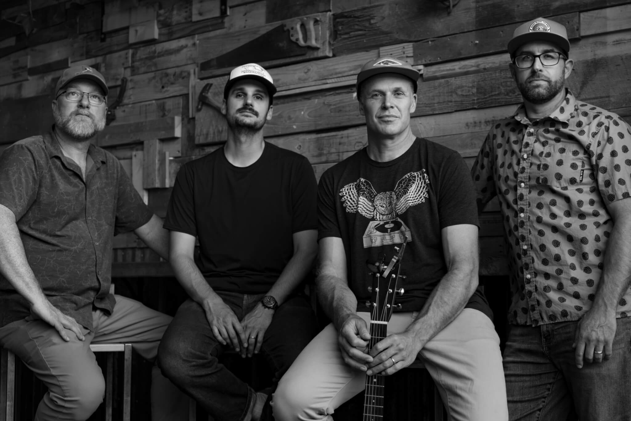 Four men in casual attire, including hats and glasses, pose together against a rustic wooden wall, with one holding an acoustic guitar in Central Oregon.