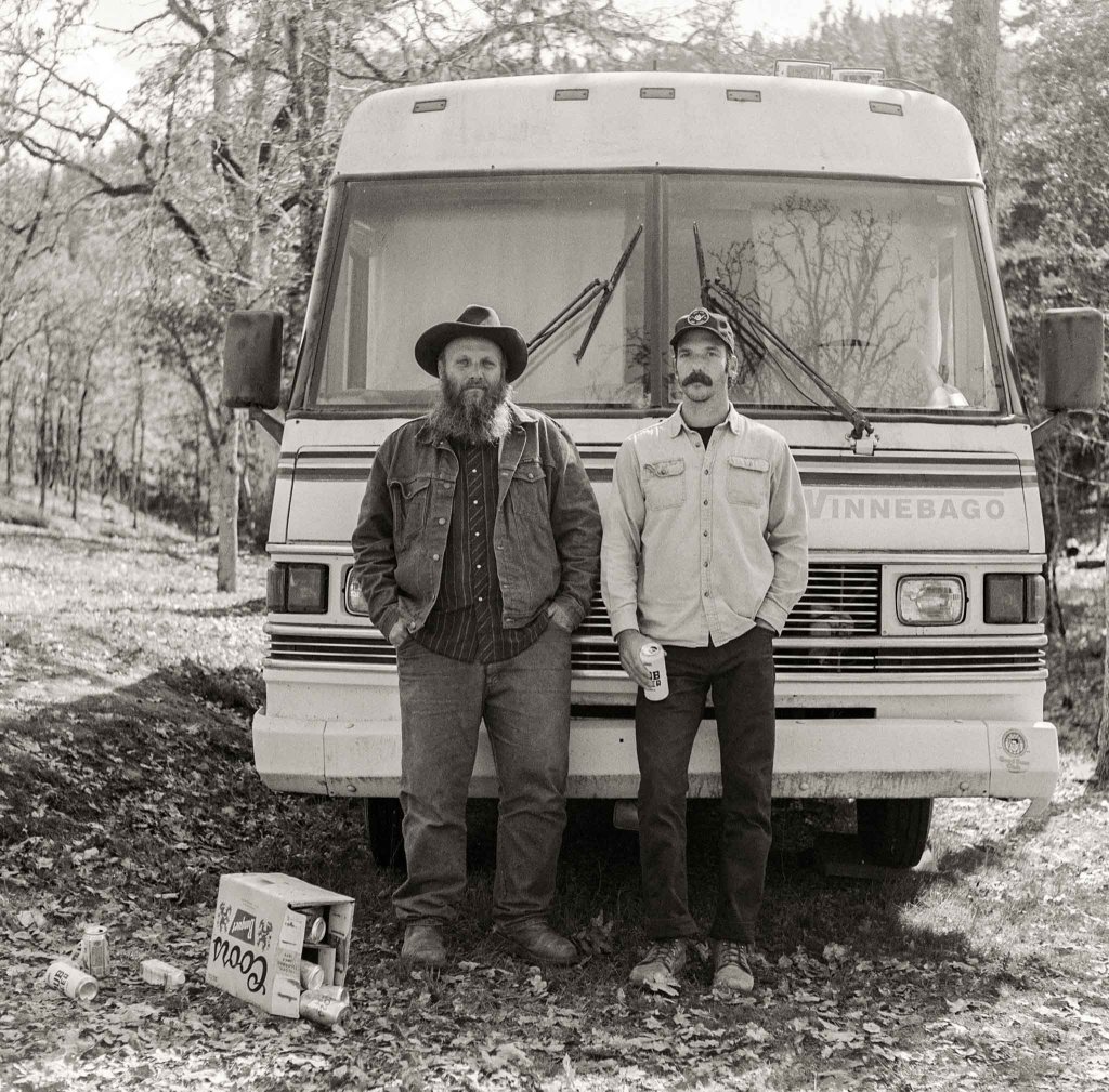 Two men stand in front of a vintage Winnebago camper van in a wooded area, surrounded by fallen leaves and beer cans in Central Oregon.