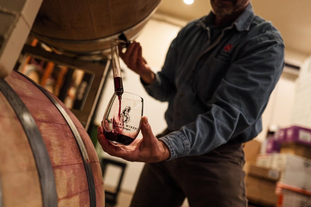 A man in a denim shirt pours red wine from a barrel into a glass with a logo, surrounded by wooden barrels in a dimly lit wine cellar in Central Oregon.