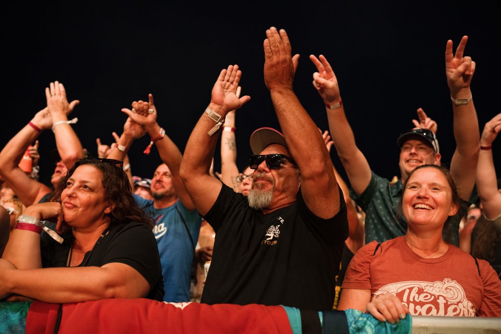 A diverse crowd of concertgoers, including a man with a beard and sunglasses, raises their hands in excitement at a nighttime outdoor event in Central Oregon.