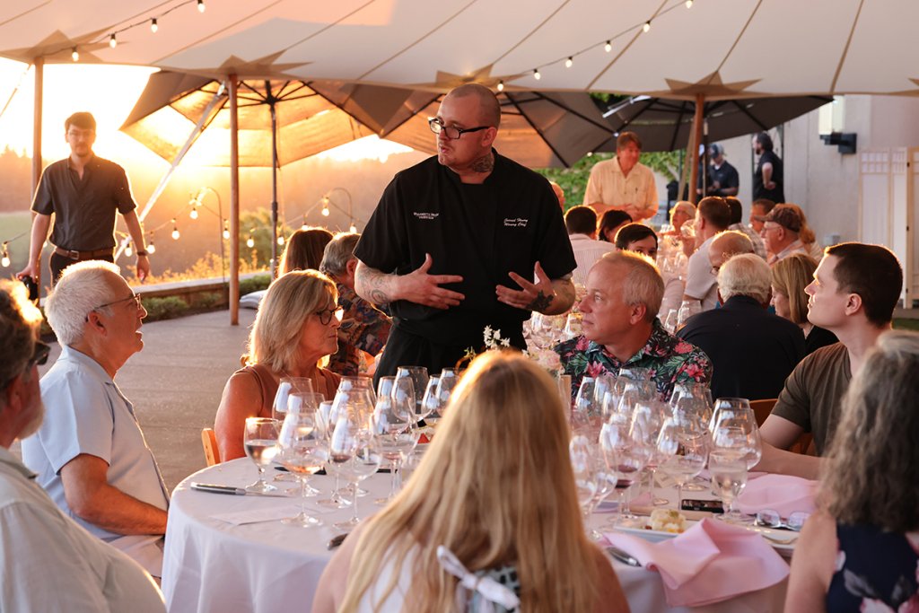 A lively outdoor dinner party under a large white tent at sunset, with guests seated at round tables adorned with wine glasses and pink napkins, and a chef addressing the crowd in Central Oregon.