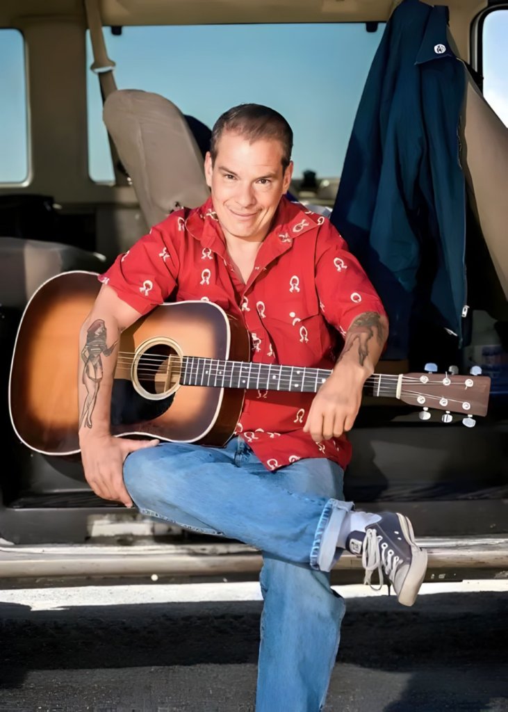 A man in a red shirt with white symbols sits on the back of a vehicle, holding an acoustic guitar and smiling in Central Oregon.
