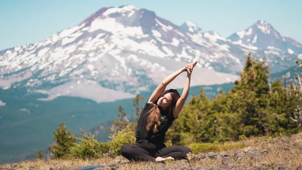 A woman performs yoga on a grassy mountain slope with snow-capped peaks in the background in Central Oregon.