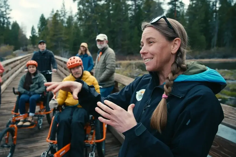 A group of people, including some in wheelchairs, enjoy a scenic bike ride on a wooden bridge surrounded by trees in Central Oregon.