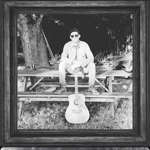 A man wearing a hat and sunglasses sits on a wooden bench with an acoustic guitar resting against his leg in Central Oregon.
