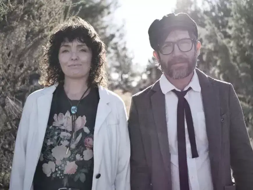 A couple stands outdoors, the woman in a floral top and white cardigan, the man in a suit with a bow tie, surrounded by trees and shrubs in Central Oregon.
