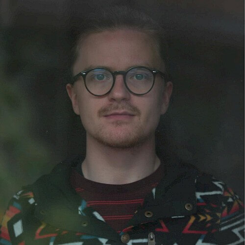 A man with glasses wearing a colorful patterned jacket stands against a dark background in Central Oregon.