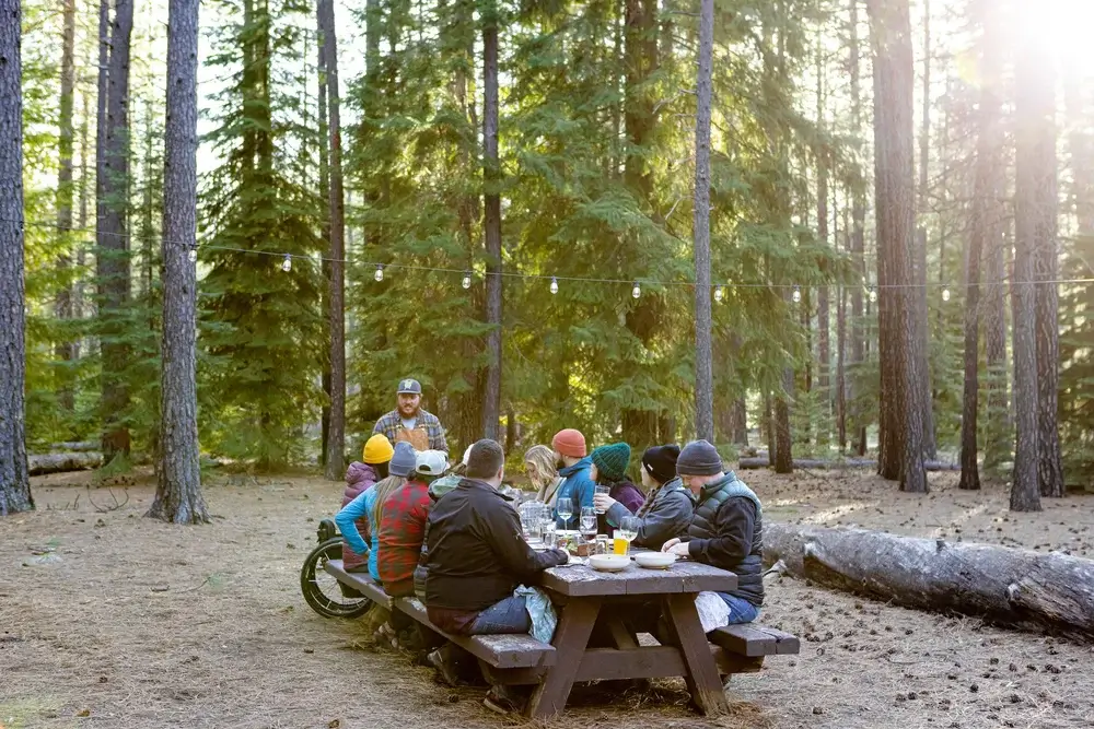 A group of people enjoys a meal together at a picnic table in a serene forest setting in Central Oregon.