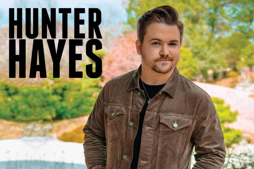 Hunter Hayes, a country music artist, stands outdoors in a stylish brown corduroy jacket against a backdrop of lush greenery and blooming trees in Central Oregon.