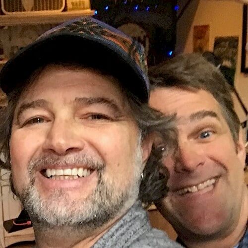 Two men smiling broadly, one wearing a patterned cap and the other with short hair in Central Oregon.