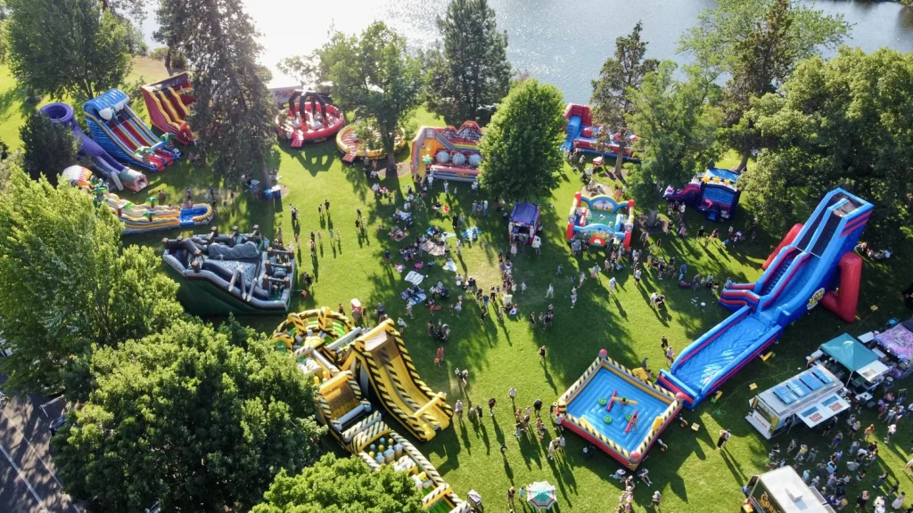 A vibrant outdoor inflatable carnival with colorful slides, bouncy castles, and games set in a lush green park by a lake, bustling with excited children and families in Central Oregon.