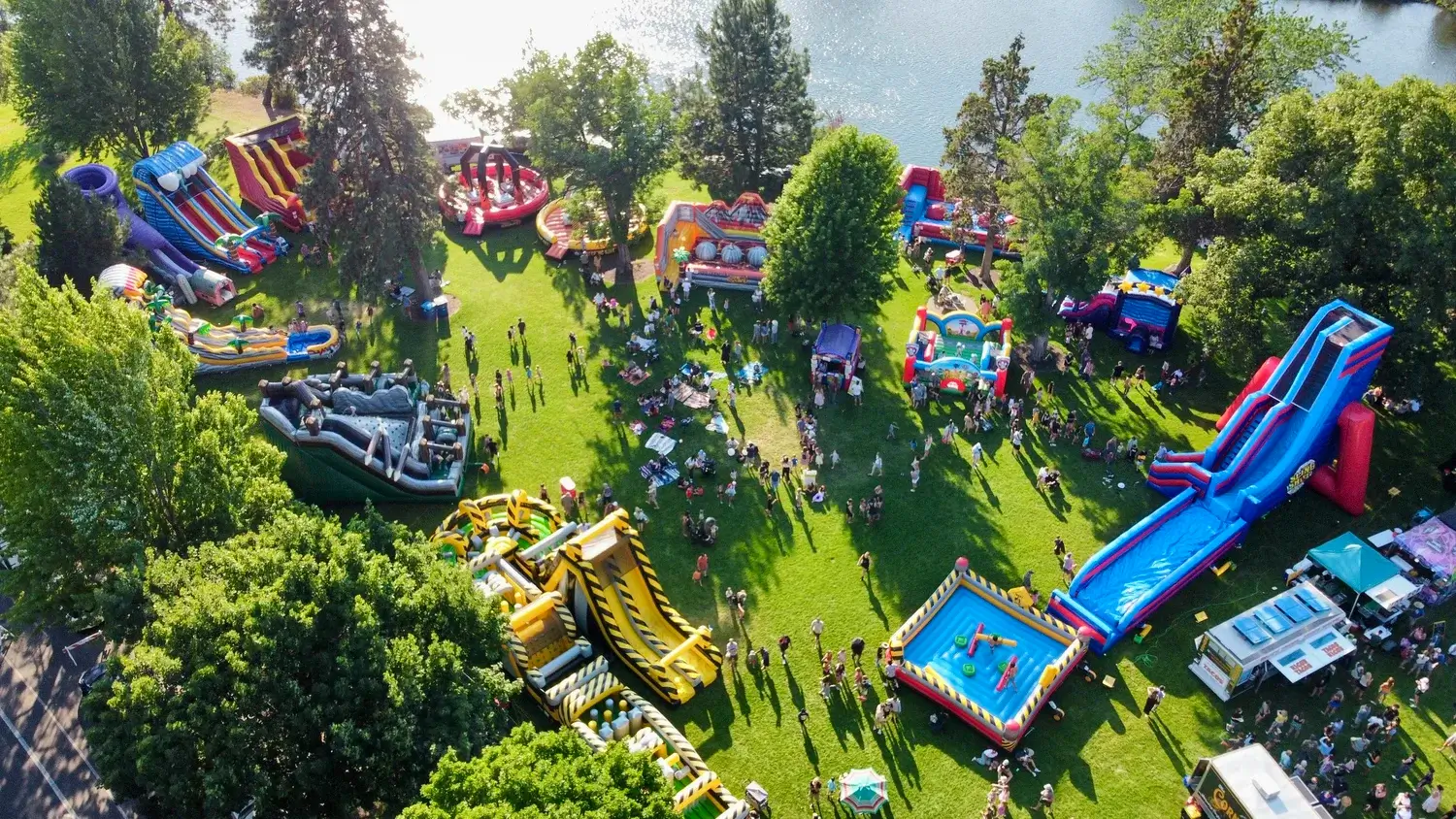 A vibrant outdoor inflatable carnival with colorful slides, bouncy castles, and games set in a lush green park by a lake, bustling with excited children and families in Central Oregon.