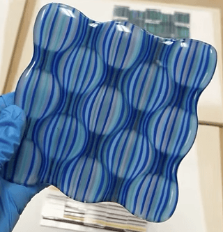 A hand wearing a blue glove holds a glass tile with a wavy, blue and white striped pattern in Central Oregon.