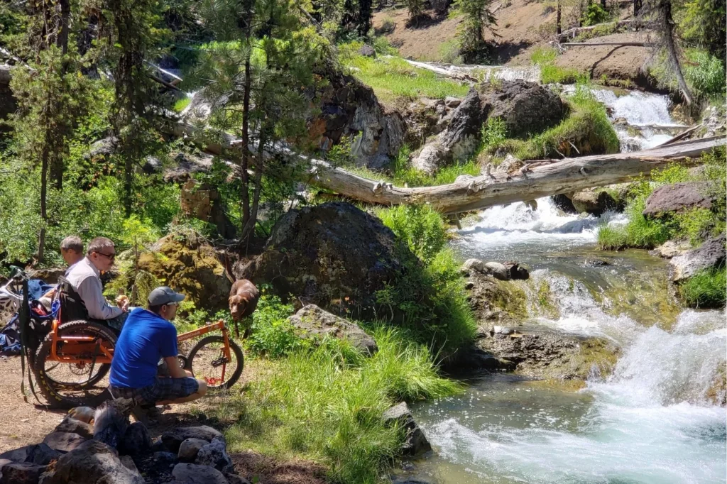 Two men in a wheelchair and on a bicycle enjoy a scenic picnic by a rushing mountain stream surrounded by lush greenery and fallen logs in Central Oregon.