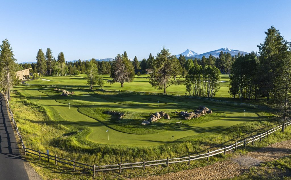A scenic golf course with rolling greens, surrounded by tall pine trees and distant snow-capped mountains under a clear blue sky in Central Oregon.