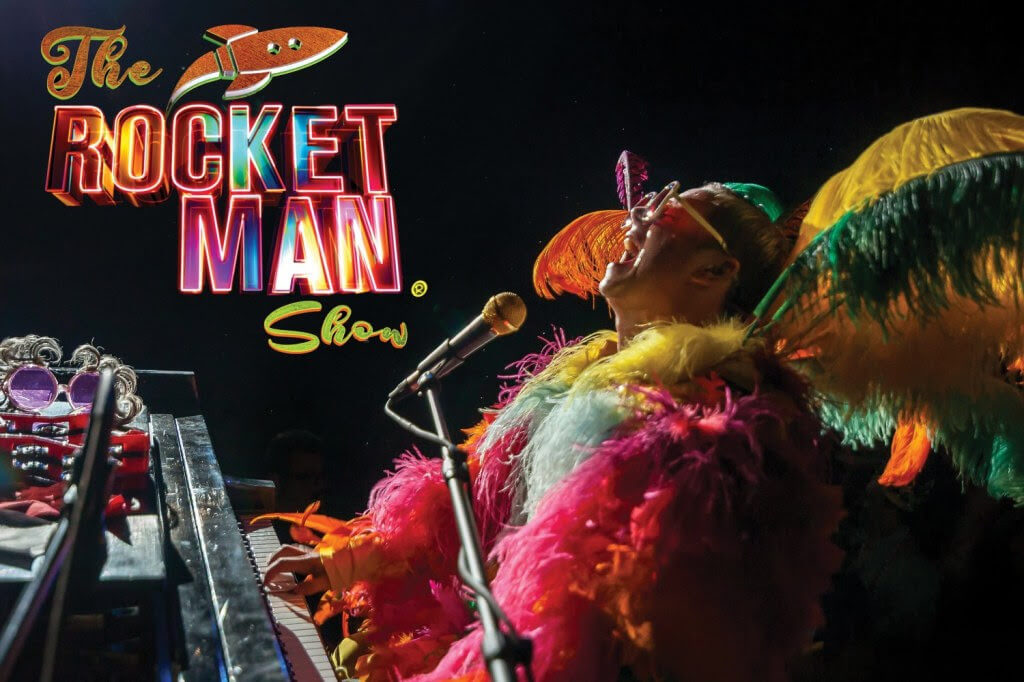 A vibrant performer in a colorful feathered costume plays piano on stage at The Rocket Man Show in Central Oregon.