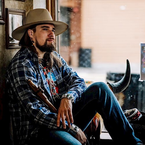 A man with long hair and a beard, wearing a cowboy hat and plaid shirt, sits with a guitar in a rustic setting in Central Oregon.