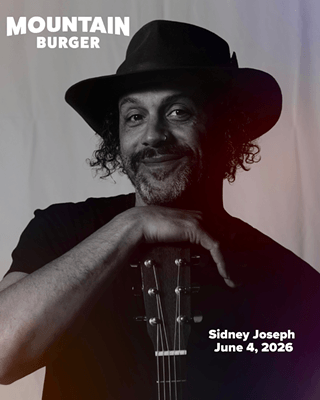A musician with curly hair and a beard, wearing a black hat and holding a guitar, smiles warmly at the camera in Central Oregon.
