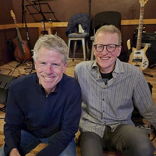 Two smiling musicians sitting in a cozy music studio filled with guitars and amplifiers in Central Oregon.