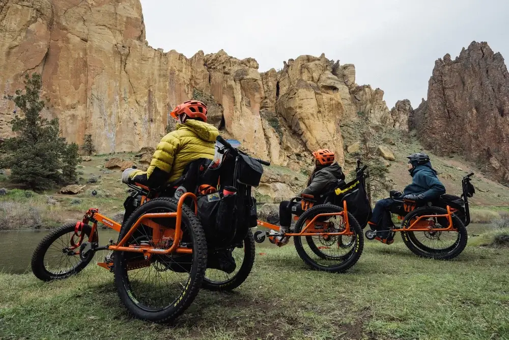 Three individuals in adaptive mountain bikes explore a scenic desert landscape with towering rock formations and a serene river in Central Oregon.