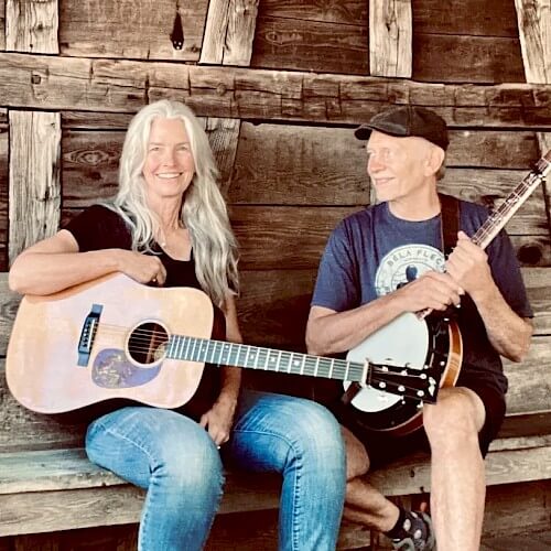 Two musicians, a woman with long gray hair and a man with a bald head, sit on a rustic wooden bench, each holding an acoustic stringed instrument in Central Oregon.