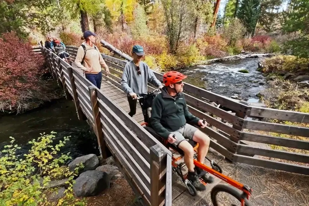 A group of people, including a person in a wheelchair, cross a wooden bridge over a rushing stream in a colorful autumn forest in Central Oregon.
