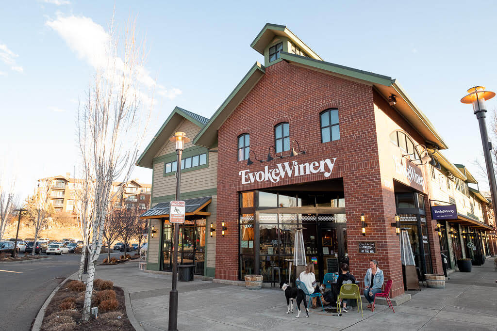 A charming brick building with a sign reading 'Evoke Winery' features outdoor seating where patrons enjoy wine with their dogs in Central Oregon.
