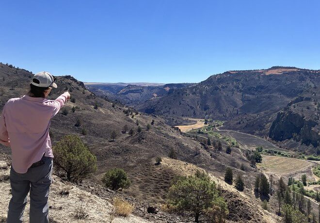 A person in a pink shirt and hat stands on a rocky hillside, pointing towards a vast canyon landscape with winding rivers and rugged mountains under a clear blue sky in Central Oregon.