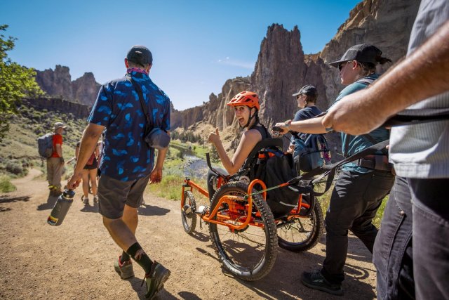 A smiling adaptive cyclist gives a thumbs-up from a hand-powered trail cycle as a team of guides assists on the trail below the Smith Rock cliffs