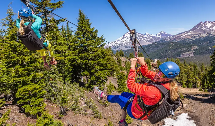 Two people ziplining through a lush forest with snow-capped mountains in the background in Central Oregon.