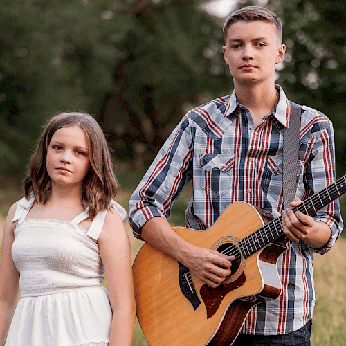 A young girl in a white dress stands beside a boy playing an acoustic guitar in a grassy field in Central Oregon.