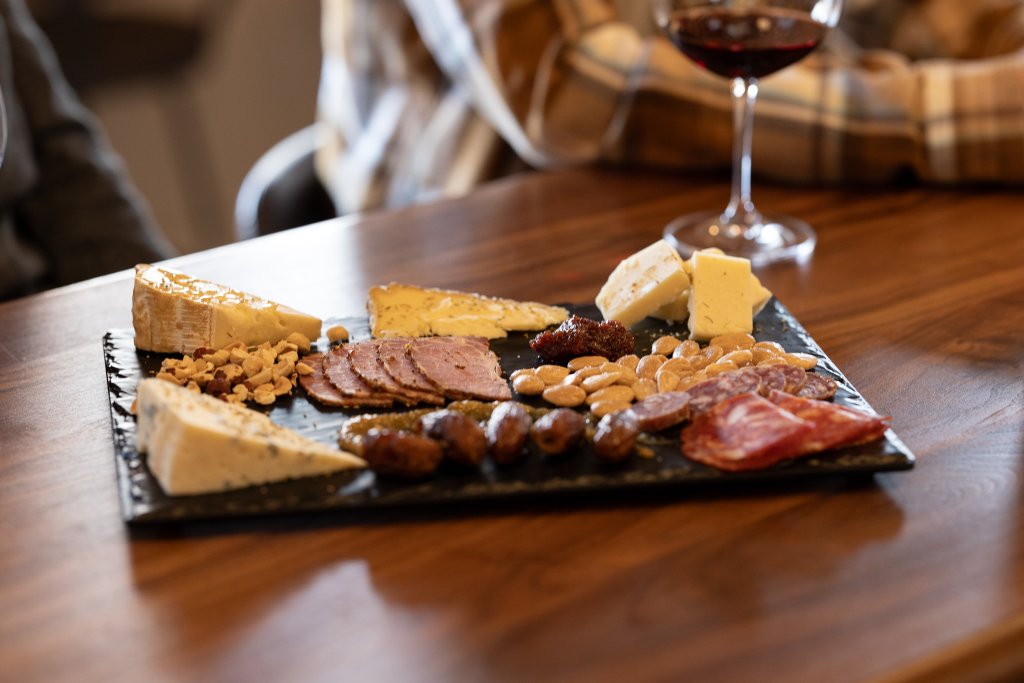 A variety of cheeses, cured meats, nuts, and dried fruits are artfully arranged on a black slate charcuterie board, accompanied by a glass of red wine in Central Oregon.