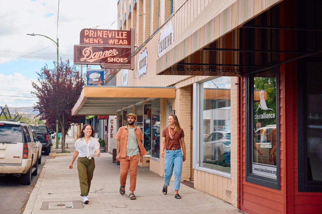 Three people stroll down a quaint small-town street lined with vintage storefronts and parked cars in Central Oregon.