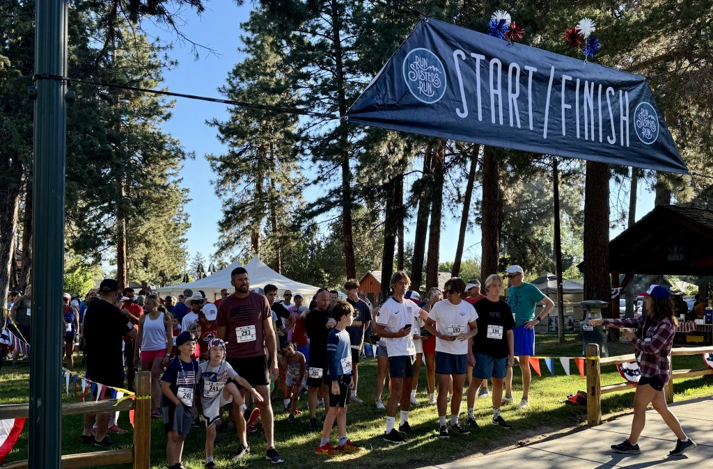 A group of runners, including children and adults, gather at the start/finish line of a race in a park surrounded by tall trees in Central Oregon.