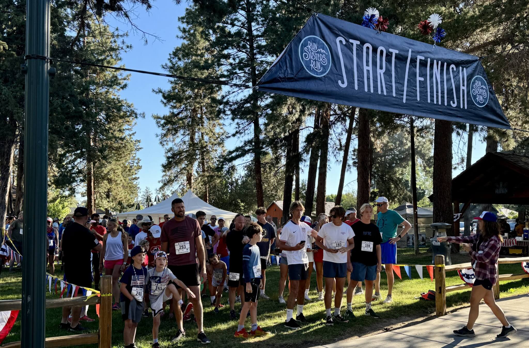 A group of runners, including children and adults, gather at the start/finish line of a race in a park surrounded by tall trees in Central Oregon.