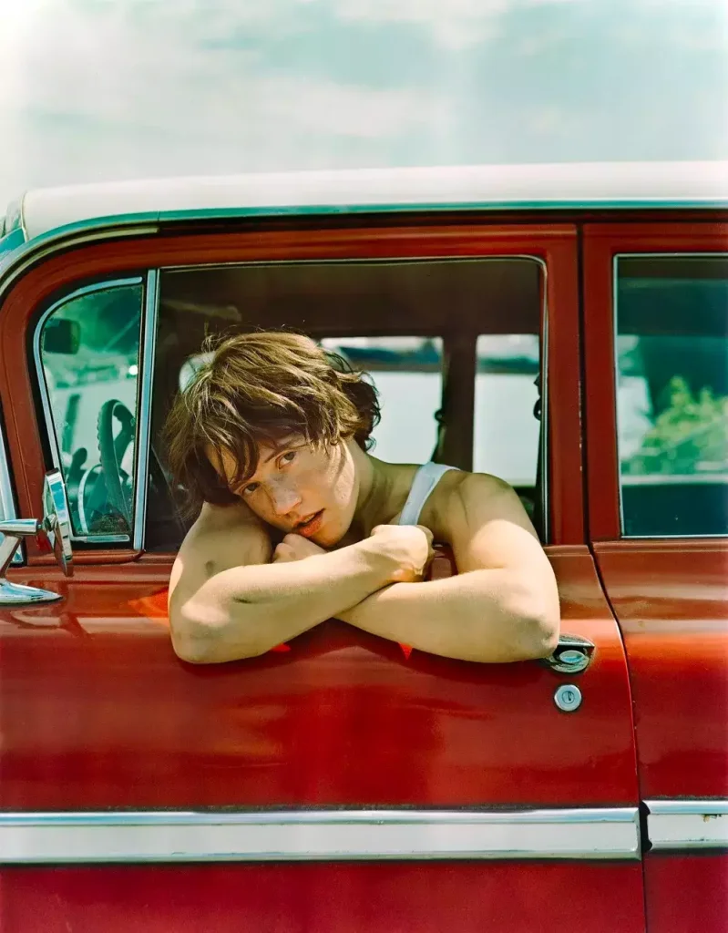 A young woman with short brown hair leans out of the window of a vintage red convertible, her arms resting on the door frame as she gazes thoughtfully into the distance in Central Oregon.