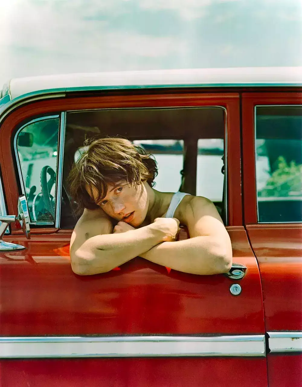 A young woman with short brown hair leans out of the window of a vintage red convertible, her arms resting on the door frame as she gazes thoughtfully into the distance in Central Oregon.