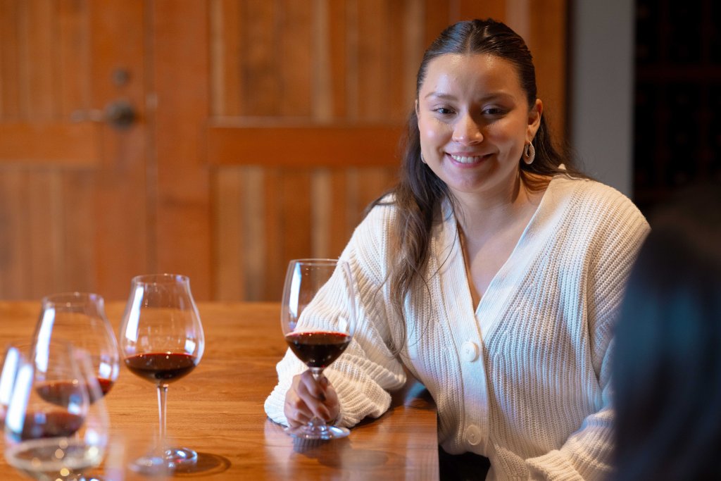A woman in a white sweater smiles while holding a glass of red wine at a wooden table with several other wine glasses in Central Oregon.